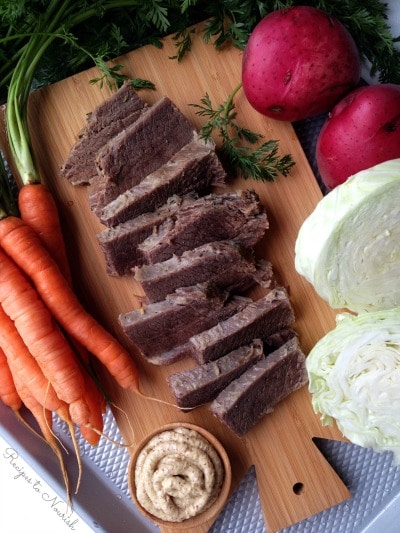 Slices of corned beef on a cutting board with carrots, cabbage, red potatoes and Dijon mustard.