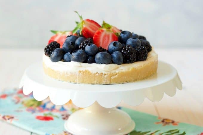 Cheesecake tart topped with fresh blueberries, strawberries and blackberries sitting on a cake stand.