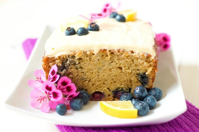 Frosted loaf cake with a slice cut off with fresh blueberries, lemon slices and geranium flowers.