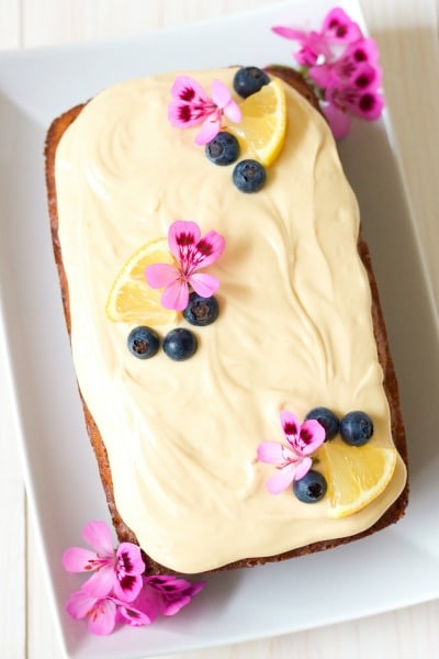 Overhead view of frosted loaf cake with fresh blueberries, lemon slices and geranium flowers.