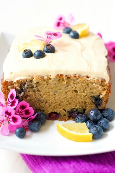 Frosted loaf cake with a slice cut off with fresh blueberries, lemon slices and geranium flowers.