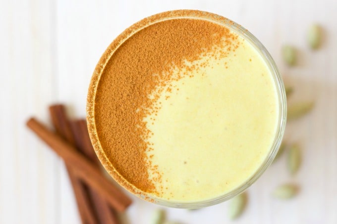 Glass of golden milk smoothie dusted with ground cinnamon, cinnamon sticks and cardamom pods.