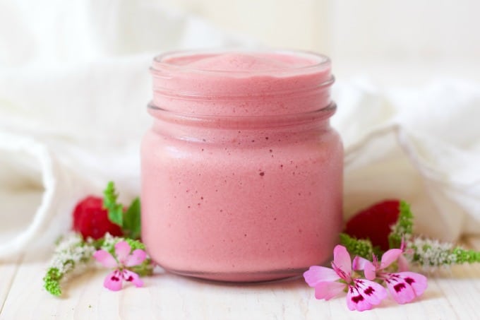 Jar of pink smoothie next to fresh raspberries and flowers.
