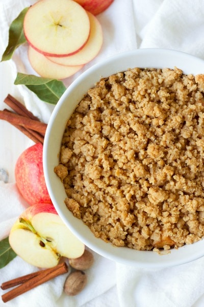 Casserole dish with apple crisp surrounded by apple slices and cinnamon sticks.