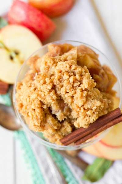 Bowl full of chunky apple crisp surrounded by apple slices and cinnamon sticks.