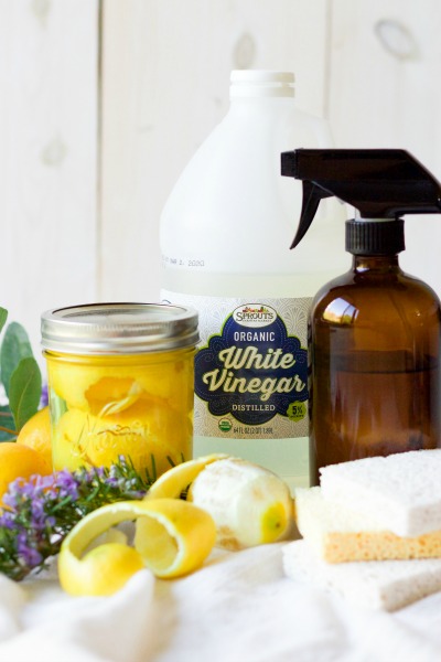 Lemon peels in a mason jar, fresh lemons, fresh herbs, kitchen dish sponges, amber glass spray bottle and Sprouts brand organic white vinegar.