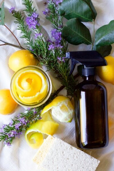 Lemon peels in a mason jar, fresh lemons, fresh rosemary and leaves, kitchen dish sponges and an amber glass spray bottle.