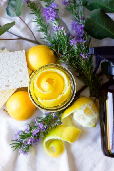 Lemon peels in a mason jar, amber glass spray bottle, fresh lemons, fresh rosemary, fresh leaves and kitchen dish sponges.