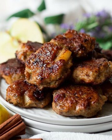 Breakfast sausage patties stacked on plates with fresh apples and cinnamon sticks.