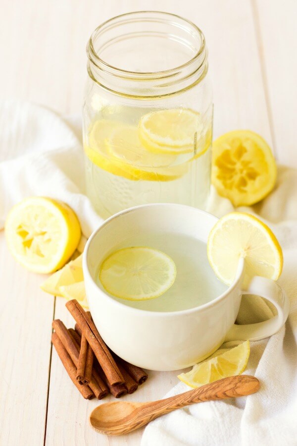 Mug and mason jar full of hot drink and lemon slices, next to lemons and cinnamon sticks.