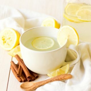 Mug and mason jar full of hot drink and lemon slices next to lemons and cinnamon sticks.