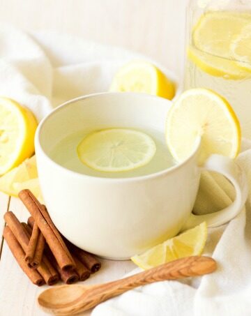 Mug and mason jar full of hot drink and lemon slices next to lemons and cinnamon sticks.