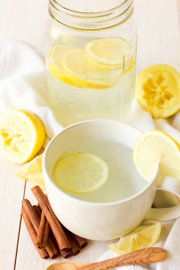 Mug and mason jar full of hot drink and lemon slices, next to lemons and cinnamon sticks.