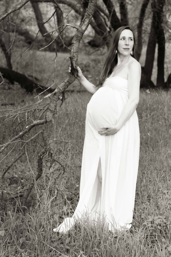 Black and white photo of a pregnant mother of twins doing a maternity photo shoot in a white maternity gown outside in nature among trees in a field.