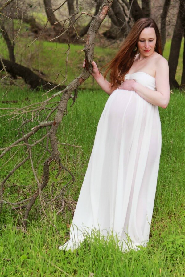 Pregnant mother of twins doing a maternity photo shoot in a white maternity gown outside in nature among trees in a field.