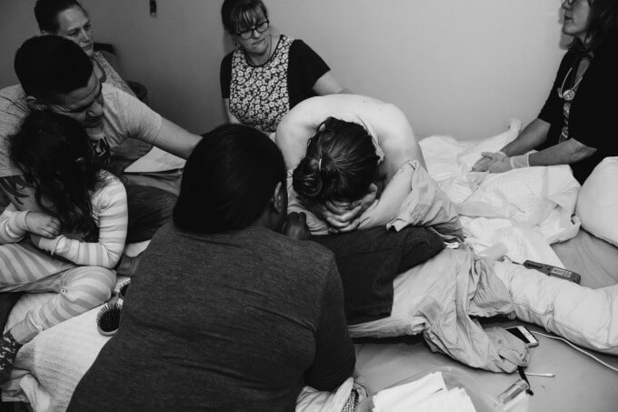 A mother in pain in active labor on her bed during a home birth surrounded by her husband and birth team.