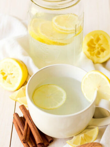 White mug filled with hot garlic ginger lemonade, topped with a lemon slice and garnished with another slice on the rim. A mason jar behind the mug is half-filled with the same drink and lemon slices, surrounded by fresh cinnamon sticks, a tiny wooden spoon, lemon wedges and squeezed lemon halves on a white wooden table with a light background.