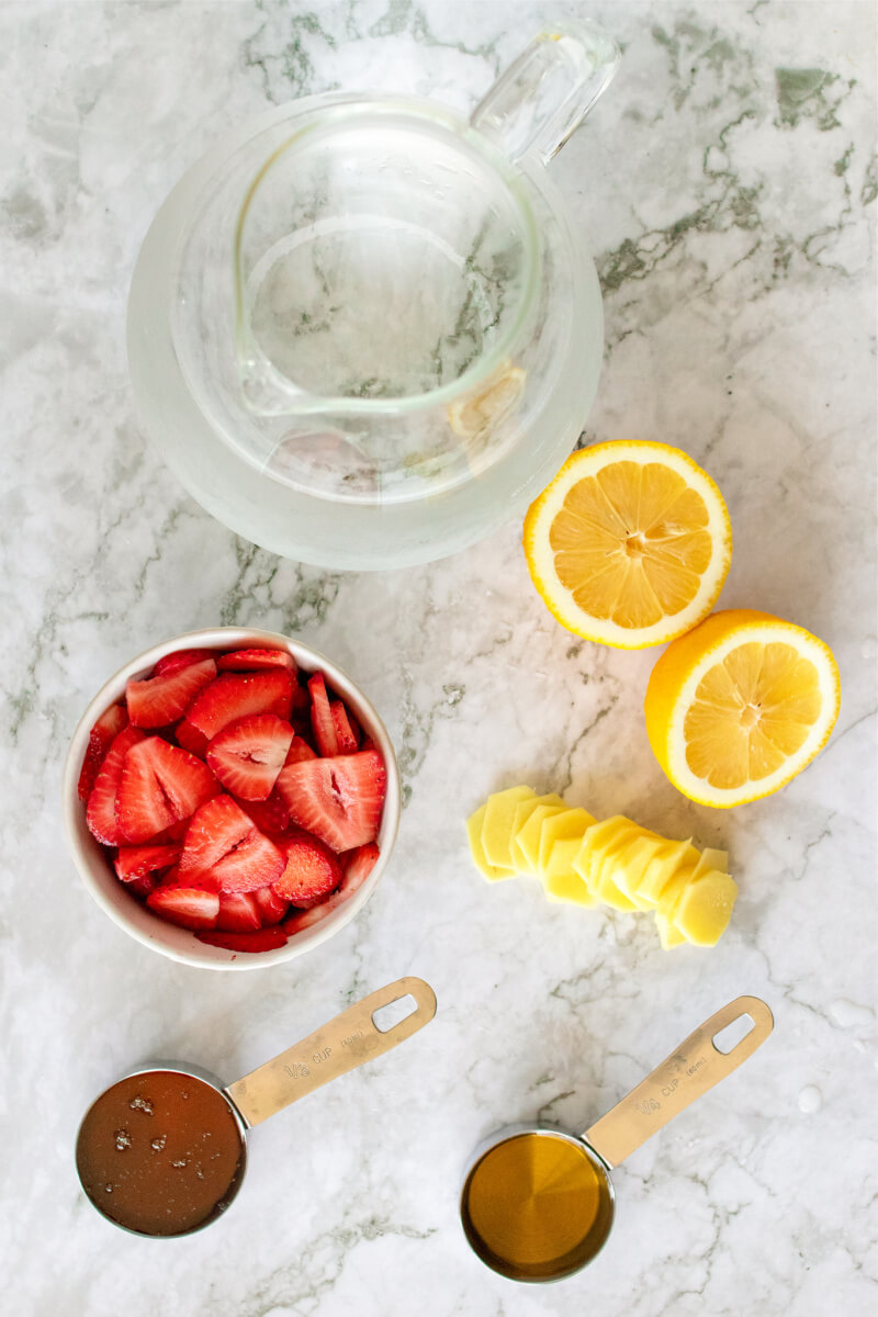 Glass pitcher filled with a water next to several ingredients - sliced fresh strawberries, sliced lemon, sliced fresh ginger and two liquid-filled measuring cups.