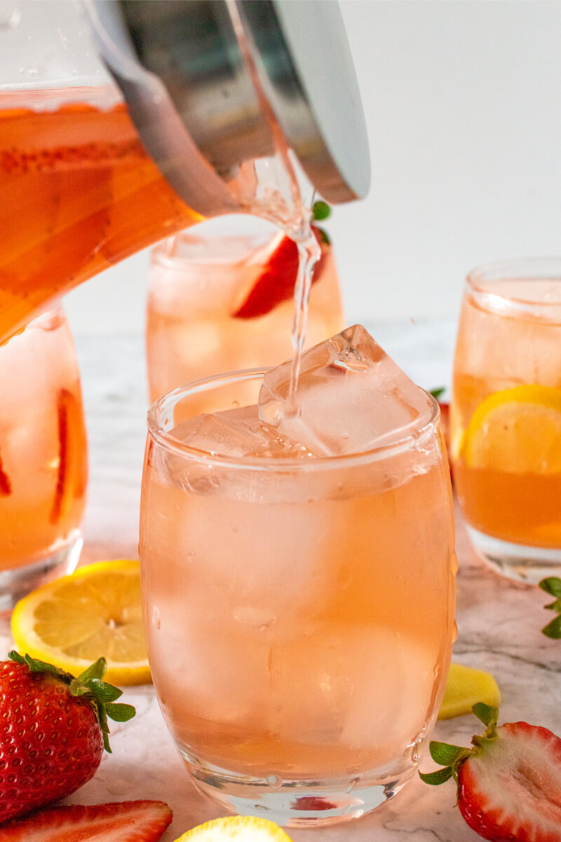 Glass pitcher pouring a strawberry drink into a glass filled with ice next to more glasses that are filled with the strawberry drink, ice, sliced lemons and sliced strawberries.