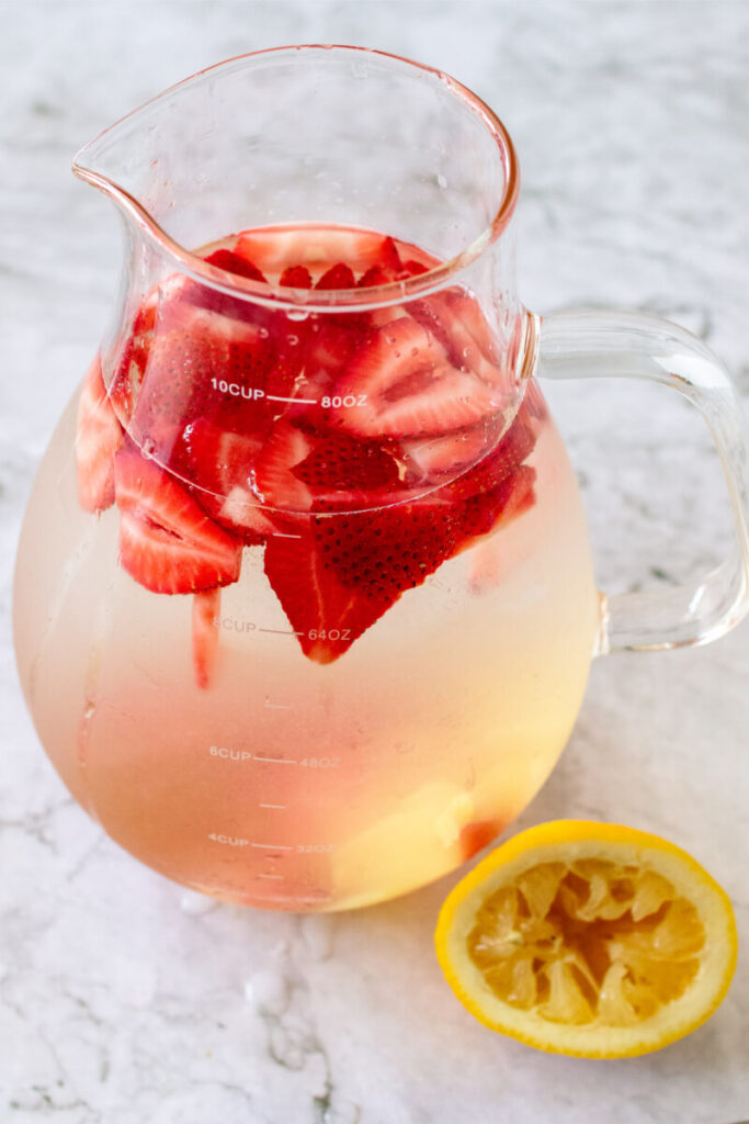 Glass pitcher filled with a water mixture and sliced fresh strawberries sitting next to a freshly squeezed lemon.