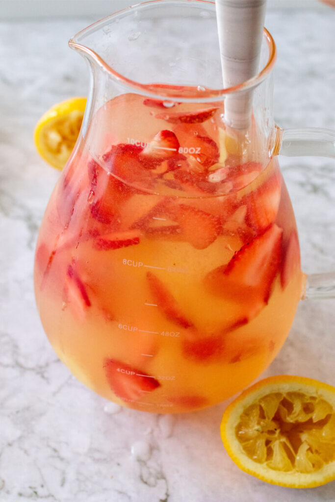 Glass pitcher filled with water mixture and sliced fresh strawberries, being stirred, sitting next to a freshly squeezed lemons.