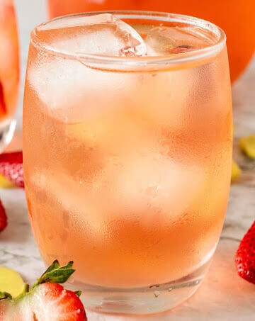 Glasses filled with ice, lemon slices, strawberry slices and a strawberry drink, sitting next to a glass pitcher filled with the drink and fresh strawberries, fresh sliced ginger and lemon slices.