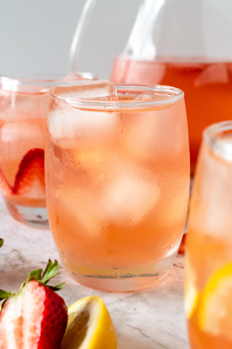 Glasses filled with ice, lemon slices, strawberry slices and a strawberry drink, sitting next to a glass pitcher filled with the drink, fresh strawberries and lemon slices.