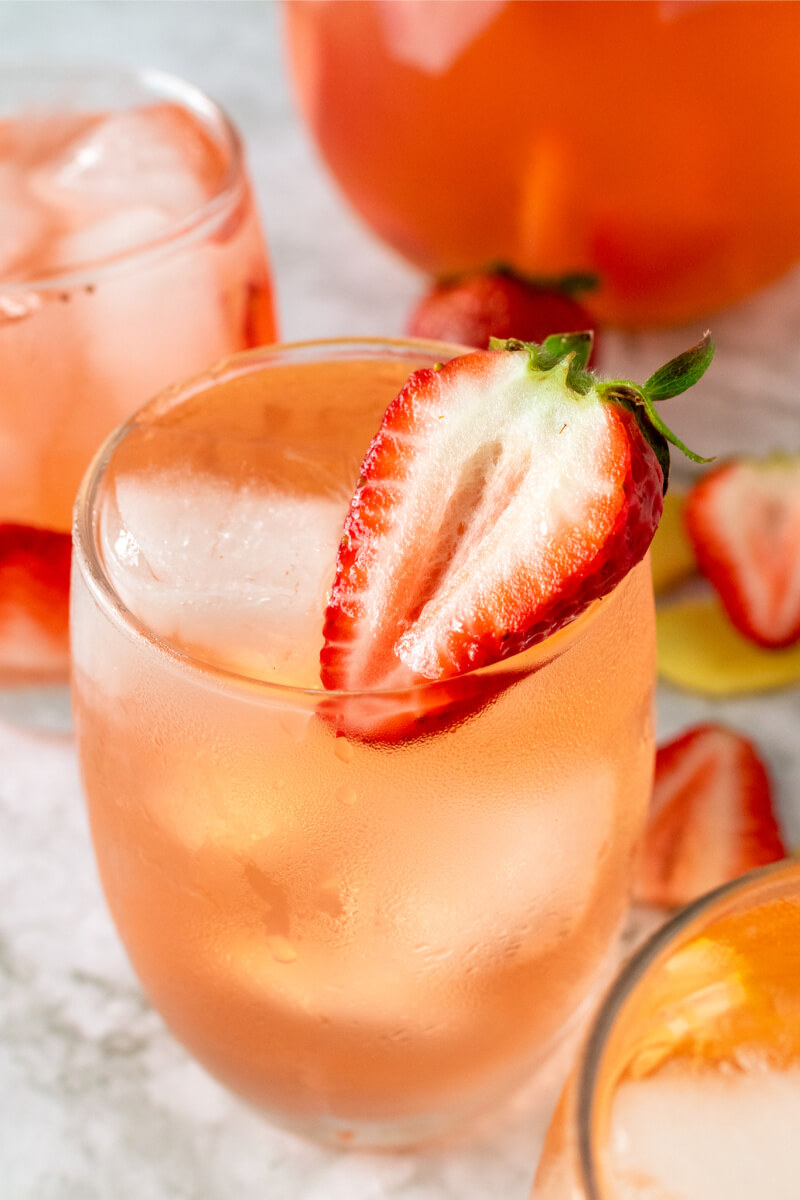 Glasses filled with ice, strawberry slices and a strawberry drink, sitting next to a glass pitcher filled with the drink, fresh strawberries and fresh sliced ginger.