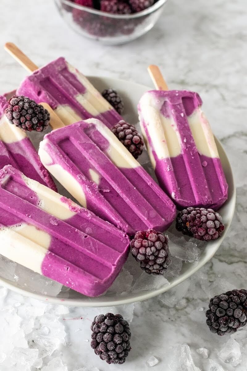 Creamy and purple swirled homemade popsicles sitting on a plate of ice next to blackberries.