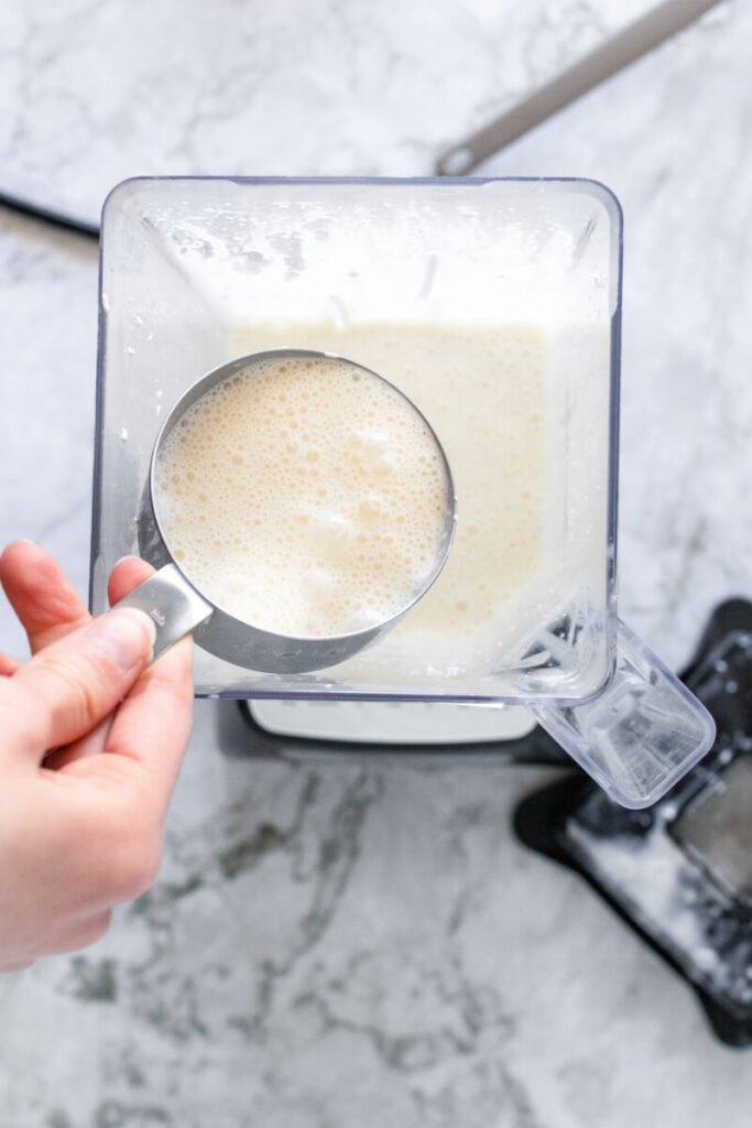 Creamy mixture in a 1-cup measuring cup being held above the mixture in a blender.