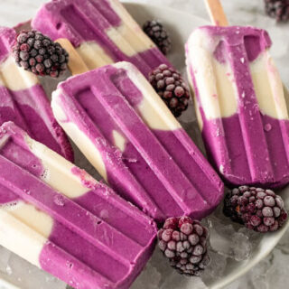 Creamy and purple swirled homemade popsicles sitting on a plate of ice next to blackberries.