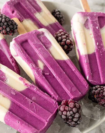 Creamy and purple swirled homemade popsicles sitting on a plate of ice next to blackberries.