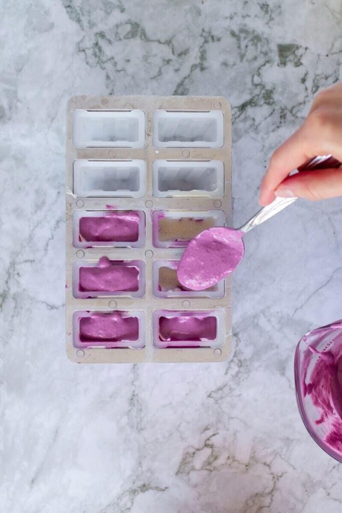 Homemade purple colored popsicles being filled and assembled in a 10-count popsicle mold next to a measuring cup of the popsicle mixture.