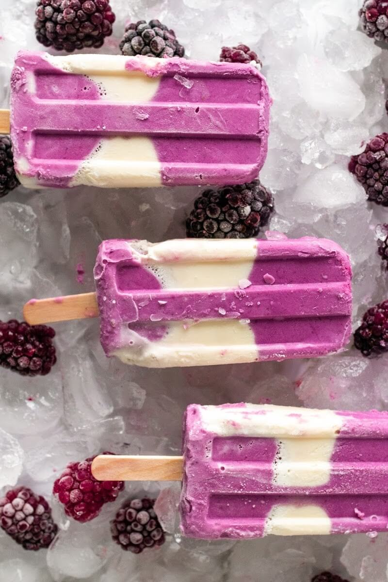 Creamy and purple swirled homemade popsicles sitting on ice next to blackberries.