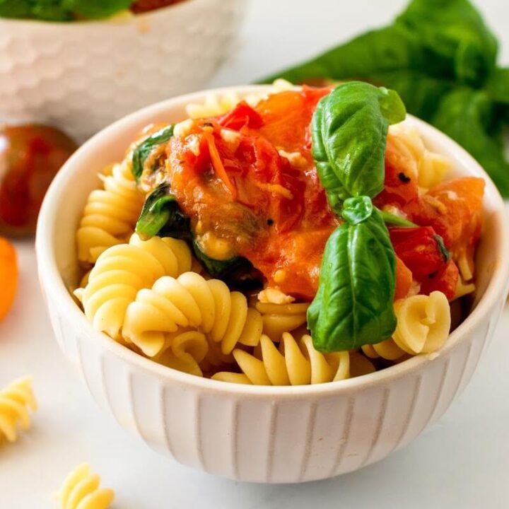Two bowls full of pasta topped with homemade tomato basil sauce with a sprig of fresh basil, surrounded by fresh cherry tomatoes, fresh basil and fresh garlic cloves.