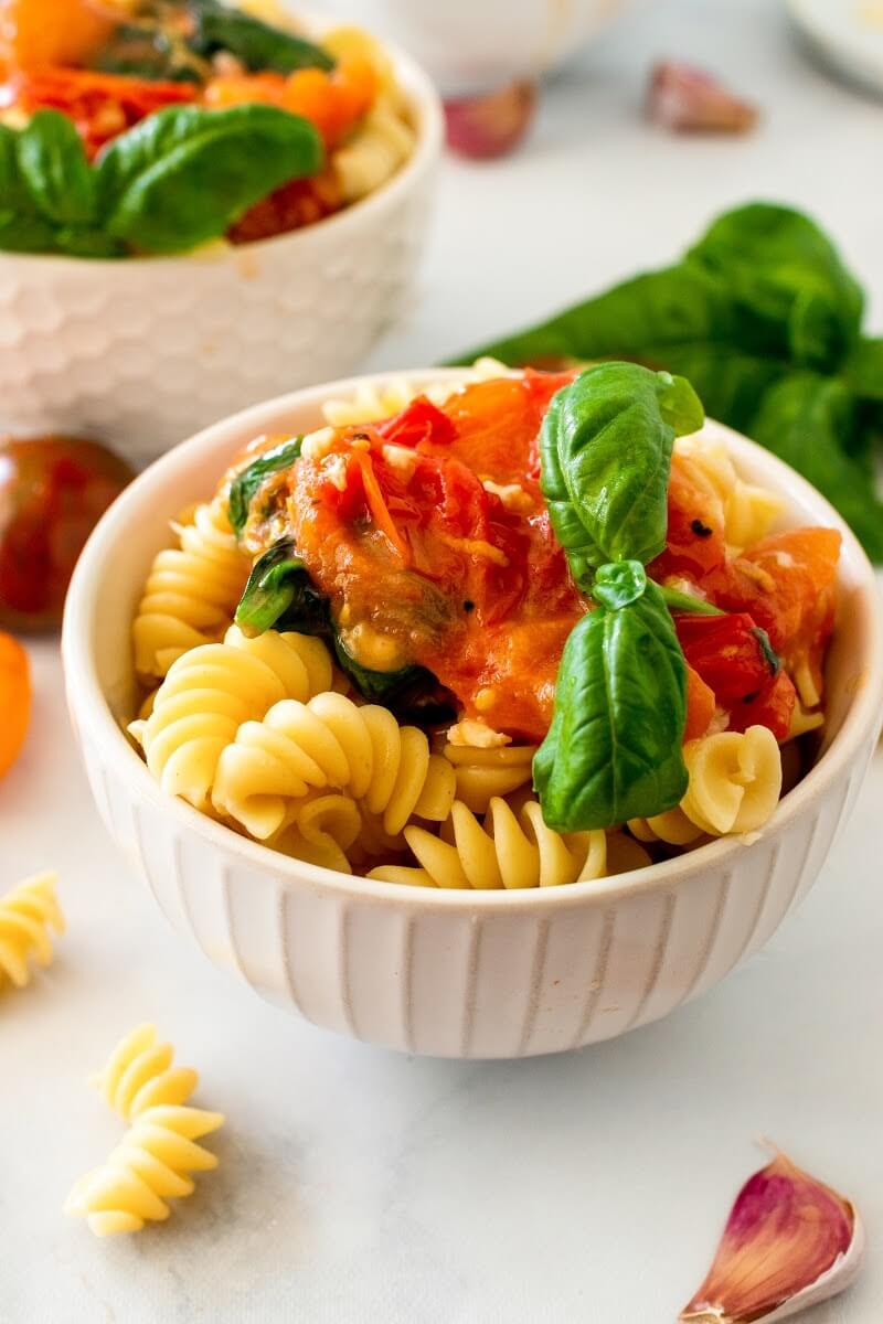 Two bowls full of pasta topped with homemade tomato basil sauce with a sprig of fresh basil, surrounded by fresh cherry tomatoes, fresh basil and fresh garlic cloves.