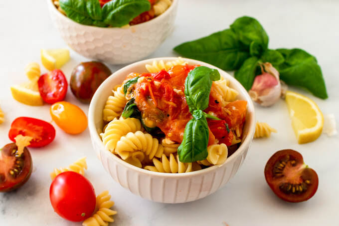 Two bowls full of pasta topped with homemade tomato basil sauce with a sprig of fresh basil, surrounded by fresh cherry tomatoes, fresh basil, lemon slices and fresh garlic clove.