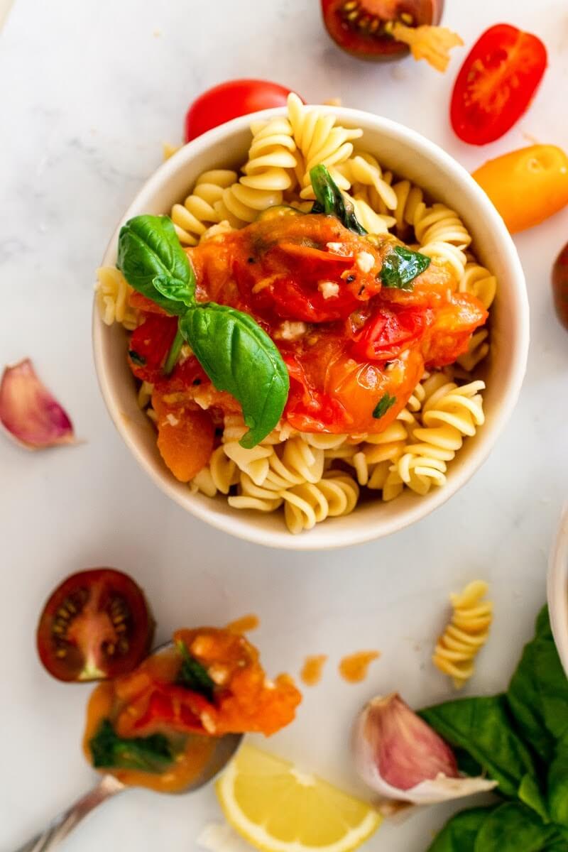 Overhead view of a bowl full of pasta topped with homemade tomato basil sauce with a sprig of fresh basil, surrounded by fresh cherry tomatoes, fresh basil, lemon slice, fresh garlic cloves and a spoonful of tomato basil sauce.