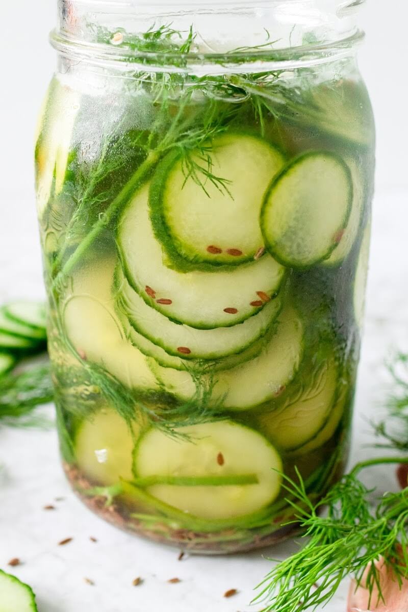 Mason jar filled with sliced pickles, fresh dill and liquid brine, surrounded by sliced cucumbers, fresh dill and spices.