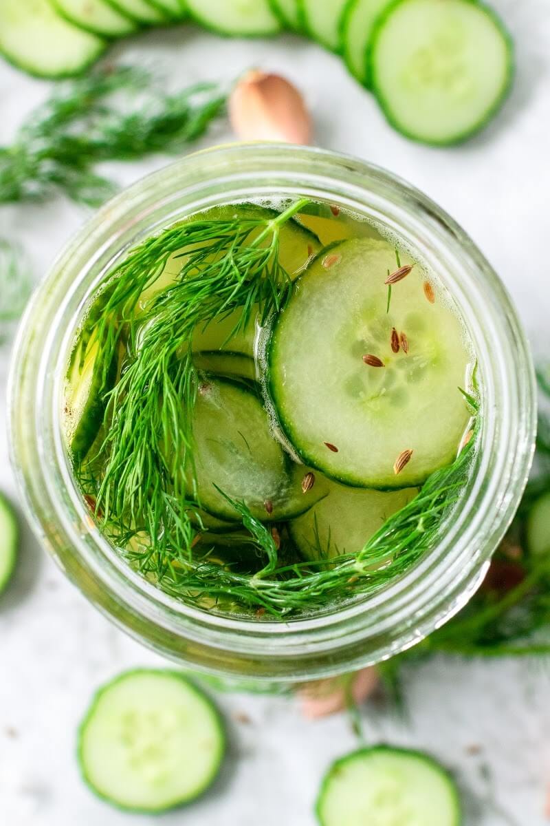Mason jar filled with sliced pickles, fresh dill and liquid brine, surrounded by sliced cucumbers, fresh dill, garlic clove and spices.