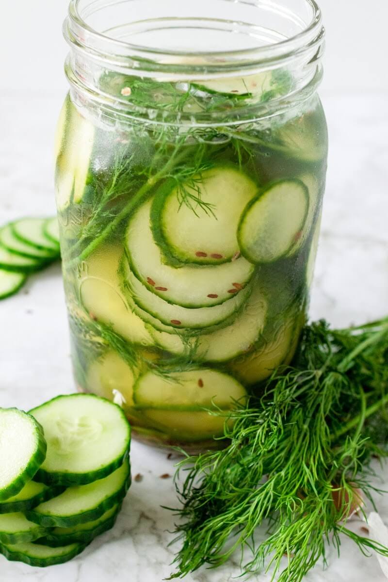 Mason jar filled with sliced pickles, fresh dill and liquid brine, surrounded by sliced cucumbers, fresh dill and spices.