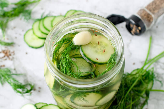 Mason jar filled with sliced pickles, fresh dill, garlic clove and liquid brine, surrounded by sliced cucumbers, fresh dill and spices.