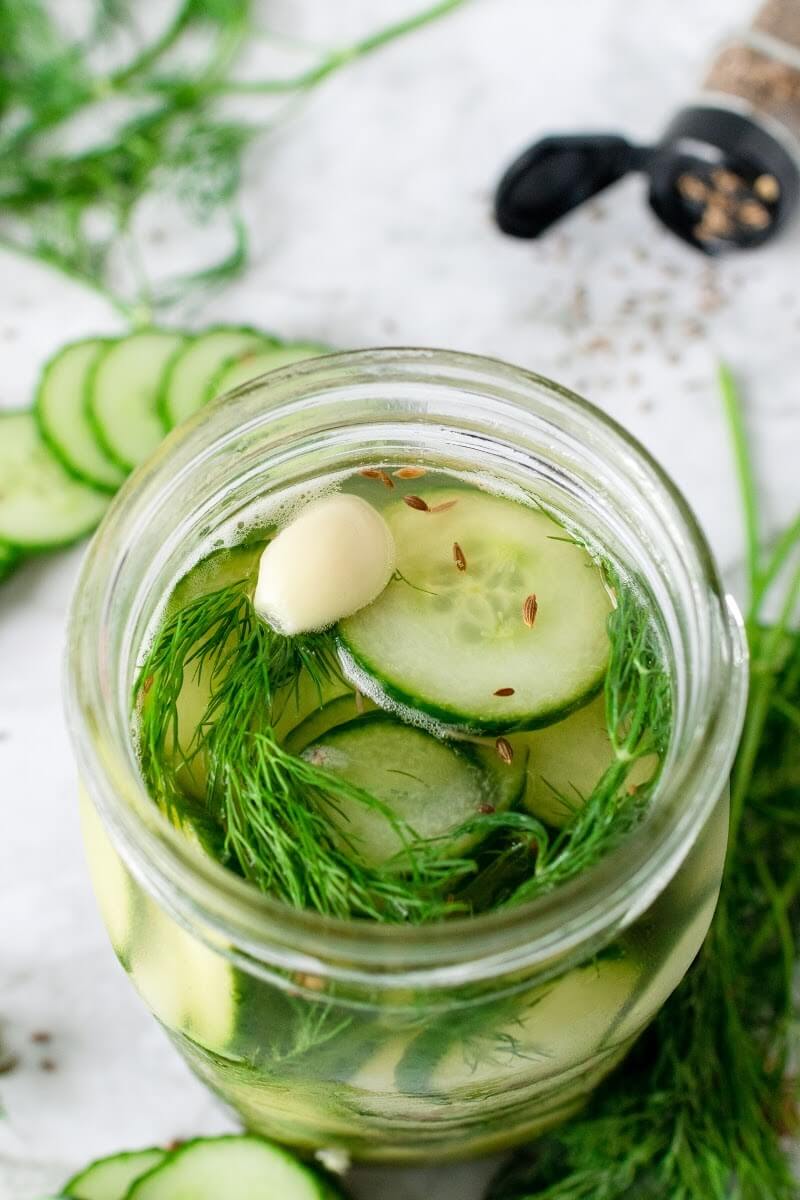 Mason jar filled with sliced pickles, fresh dill, garlic clove and liquid brine, surrounded by sliced cucumbers, fresh dill and spices.