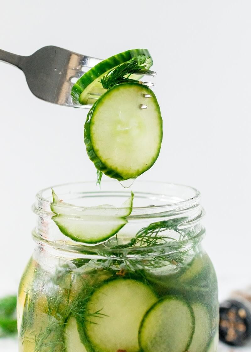 Side profile of a mason jar filled with sliced pickles, fresh dill and liquid brine, with a fork holding up two slices of pickles on the fork tines above the opening of the jar.