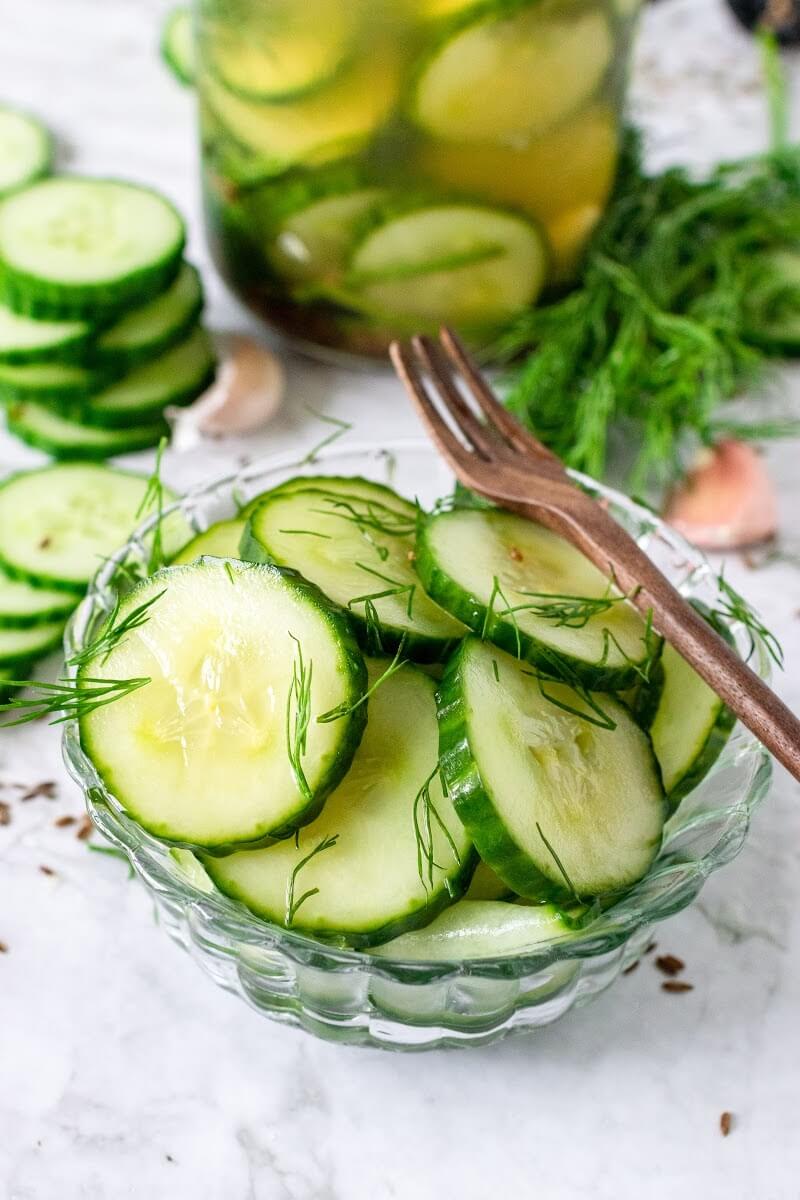 Bowl of sliced pickles with tiny fresh dill pieces on top of the pickles and a wooden fork sitting on the bowl. The background has a mason jar filled with sliced pickles surrounded by sliced cucumbers, garlic cloves and fresh dill.