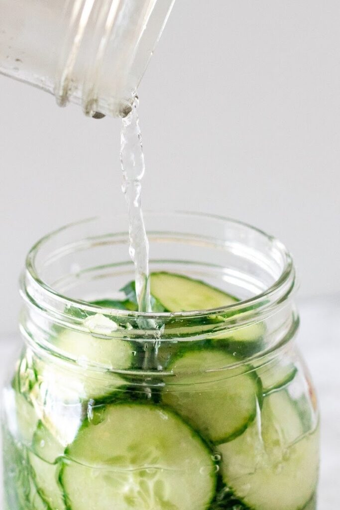 Glass jar pouring water into a mason jar filled with sliced cucumbers.