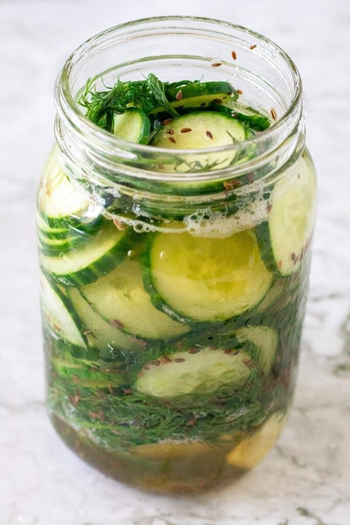 Side profile of a mason jar filled with sliced pickles, fresh dill, spices and liquid brine.