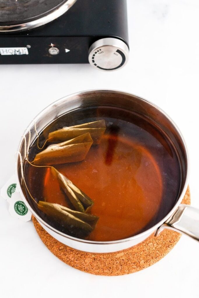 A saucepan filled with hot tea with 4 tea bags steeping in it, sitting on top of a trivet, next to an electric stovetop.