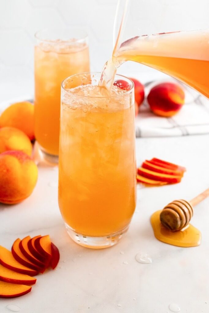 A glass full with iced peach tea and a glass pitcher filled with peach tea pouring into the glass, sitting next to sliced peaches, fresh whole peaches, a honey dipper and a glass full of iced peach tea.