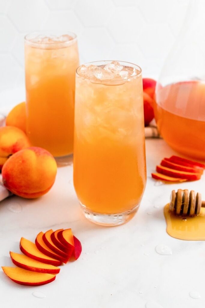 Two glasses full of iced peach tea sitting next to sliced peaches, fresh whole peaches, a honey dipper and a pitcher half full of peach tea.